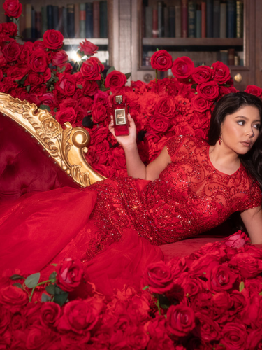 Daisy Marquez lounges on a red velvet couch, surrounded by roses, elegantly posing with her Scarlet Eau de Parfum by Bella Perversa, 2.5 fl oz.